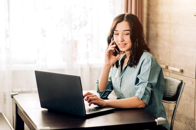 Attractive woman having happy look while speaking over smartphone, using black laptop. People, technology, lifestyle, working at home. Business woman in casual clothes consulting customer on phone