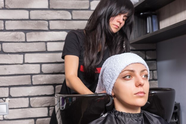 Attractive woman having her hair wrapped in a towel by female professional stylist in washing sink in beauty salon