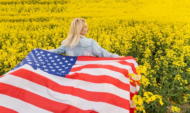 привлекательная женщина, держащая американский флаг на ветру в поле рапса. Летний пейзаж на фоне голубого неба. Горизонтальная ориентация.