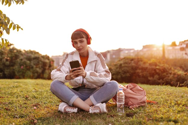 Attractive woman in jeans, white jacket sits in grass outside, holds smartphone and listens to music in red headphones