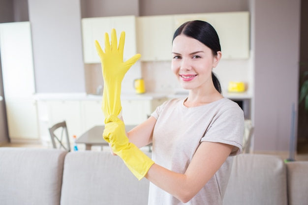 Attractive woman looks on camera. She stands in studio apartment and puts on yellow glove on right hand. Girl is smiling. She looks happy.