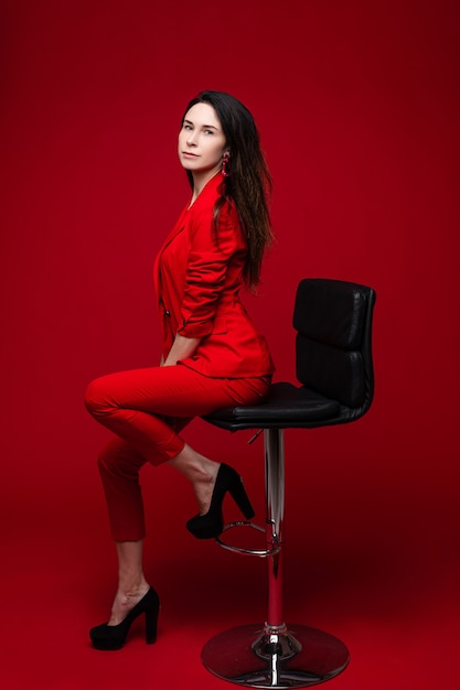 Attractive woman poses for the office magazine and sits on the chair, picture isolated on red wall