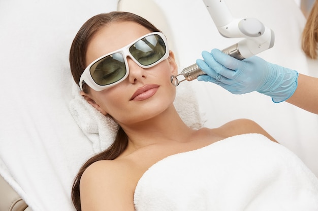 Attractive woman receiving face treatment on her cheek with laser in glasses, beautician doing facial procedures for female in spa salon