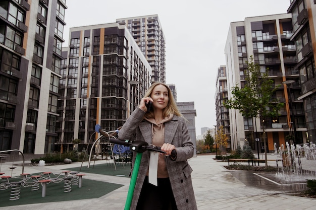 Attractive woman rides scooter and tells her friend about the benefits of renting an electric vehicle. Electrical scooter. Talking on the smartphone. Modern apartment blocks on background.