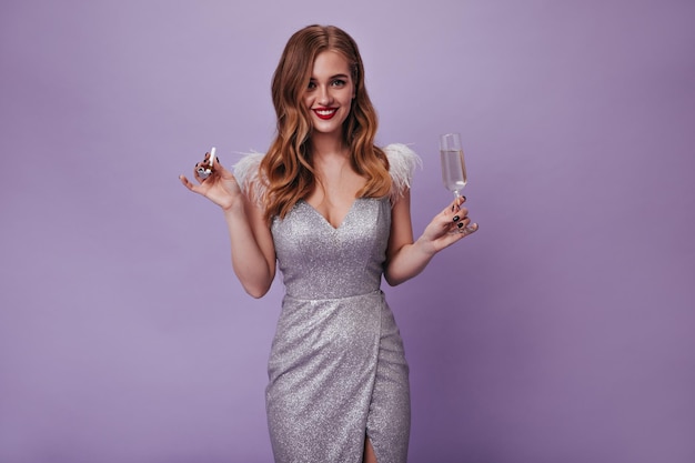 Attractive woman in silver dress looks into camera and holds champagne glass on purple background Portrait of lady in shiny outfit posing on isolated backdrop