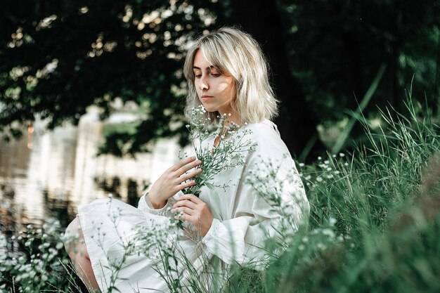 Attractive woman sits on the bank of the river on a warm day among white flowers.