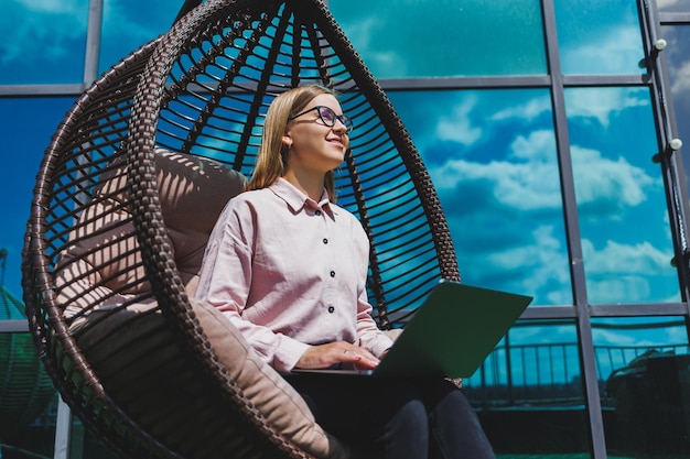 Attractive woman sits with a laptop on the balcony and works looking intently at the screen A beautiful freelancer girl in casual clothes works at a laptop while sitting in a chair on the balcony