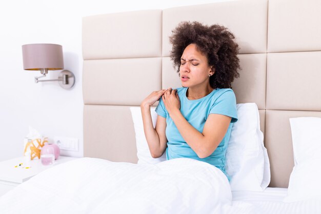 Attractive woman sitting on the bed and holding painful shoulder with another hand.