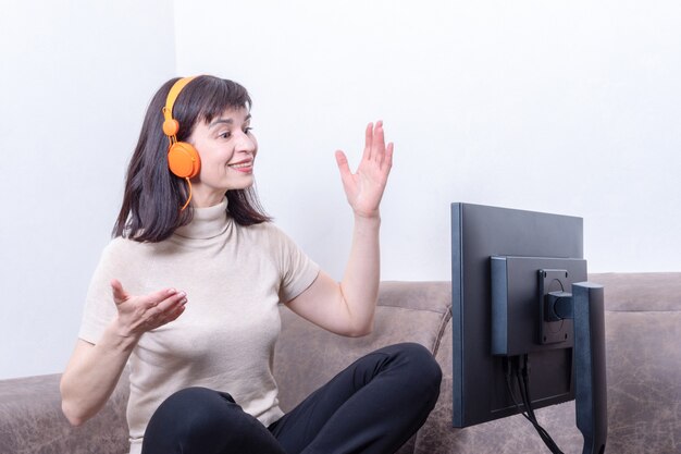 Attractive woman sitting on a sofa wearing orange headphones looking at a computer monitor, gesturing and communicating online