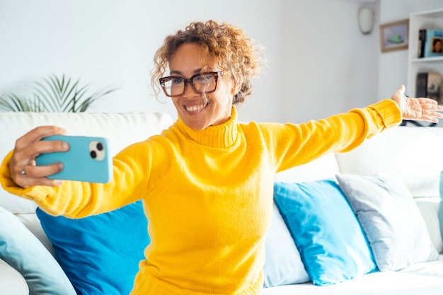 Photo attractive woman sitting on white sofa at home smiles takes selfie with her cell phone happy female with yellow sweater spends good time relaxing at home leisure and relaxation concept