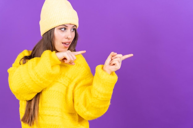 紫色の背景に黄色のウール セーターのコピー スペースで指を指して笑顔の魅力的な女性
