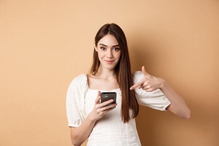 फोटो attractive woman smiling and pointing at smartphone, showing online offer on phone, standing on beige background.