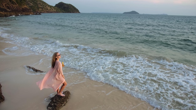 魅力的な女性は黄金の夕日の海の岩石の上に立つ緑の水着で熱帯のビーチの女の子と風に羽ばたきピンクのチュニックシルクシャツケープは彼女のオレンジ色のカクテルピニャコラーダを飲みます