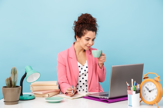 attractive woman using laptop and drinking coffee while working at desk isolated over blue wall