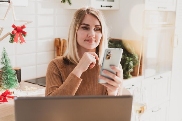 Attractive woman video chatting on phone while sitting in kitchen in festive christmas interior