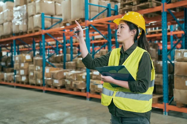 Attractive woman in warehouse checking inventory levels of\
goods on shelf. lady worker in hard hat and safety vest point\
finger looking up counting parcels and cardboard boxes in large\
storehouse.