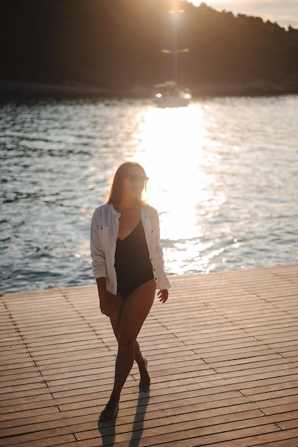 Attractive woman wearing black swimming suit and white shirt standing on pier with crossed legs and looking aside Positive lady in sunglasses posing on seascape background with yacht