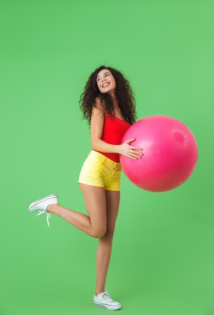 attractive woman wearing summer clothes doing exercises with fitness ball during aerobics against green wall