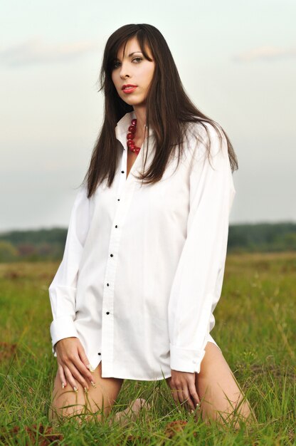 Attractive woman in white shirt with red accessories posing in field sitting on grass. Outdoor portrait