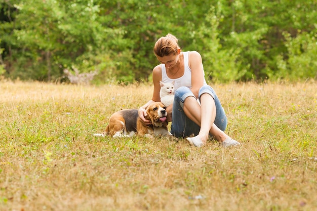 Attractive woman in a white top and blue jeans sitting on grass, embracing white cat, looking at her dog, laying near. Green forest background