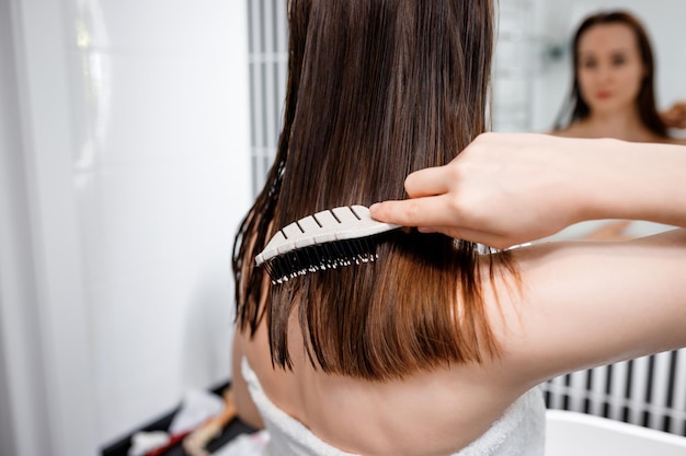 Attractive woman in white towel with comb brushing her wet hair\
after showering at home in front of bathroom mirror cares about\
healthy and clean hair beauty concept