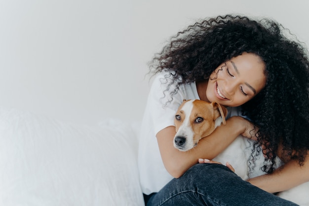 Attractive woman with curly Afro hairstyle, cuddles and pets dog with smile, expresses love, enjoys cozy domestic atmosphere