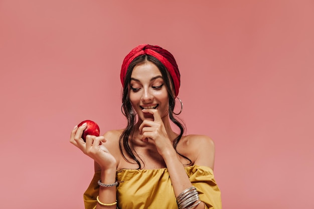 Foto donna attraente con capelli ricci in elegante bandana luminosa e orecchini rotondi guardando mela rossa su sfondo isolato