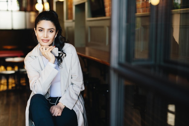 Donna attraente con capelli scuri e manicure rossa che indossa eleganti abiti bianchi seduta in un accogliente caffè godendo di buoni brani utilizzando l'app di musica gratuita sul suo smartphone. concetto di persone, tempo libero e stile di vita