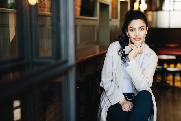 Attractive woman with dark hair and red manicure wearing stylish white clothes sitting in cozy cafe enjoying good tracks