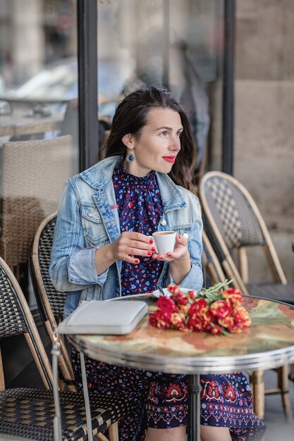 Attractive woman with long brunette hair in a blue dress sitting alone with flowers in the street cafe and drinking coffee