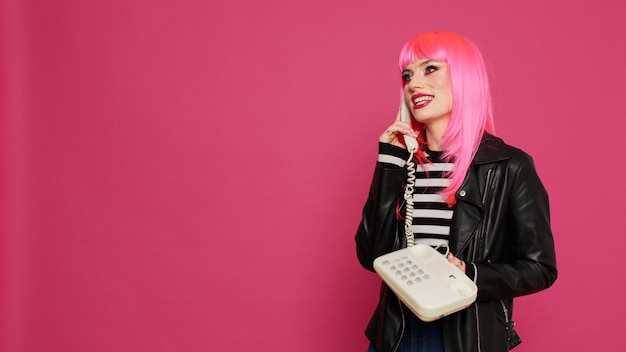 Attractive woman with pink wig talking on landline phone call,\
chatting on retro telephone line with cord in studio. having fun\
using vintage phone for remote communication, cool person.