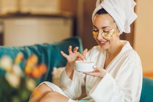 Foto una donna attraente con la cura della pelle sotto le bende sugli occhi che applica una crema idratante mentre si gode la mattinata a casa sua.