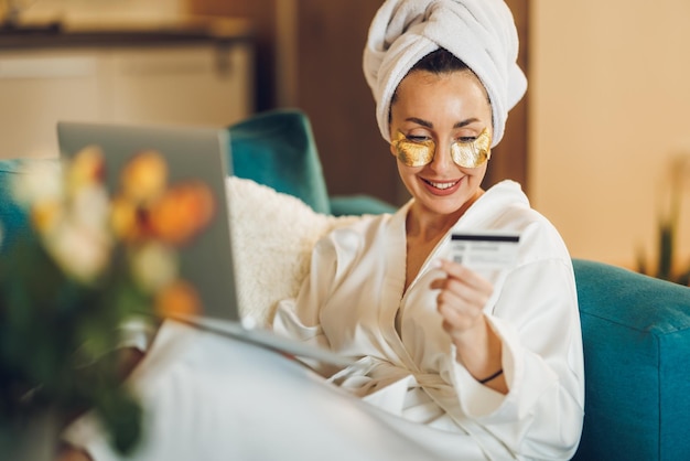 An attractive woman with skincare under eye patches shopping online on her laptop while enjoying morning at her home.