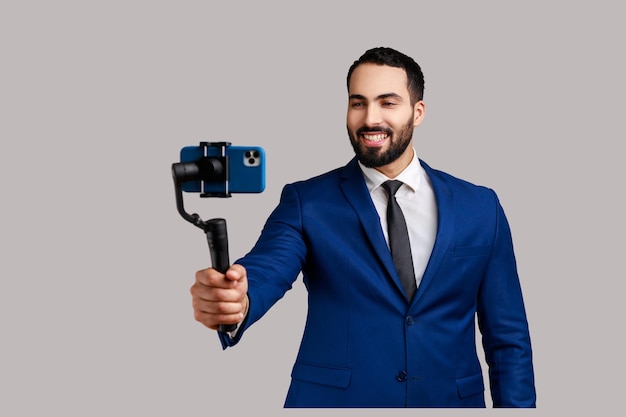 Привлекательный молодой взрослый бородатый мужчина-блогер держит стедикам с телефоном, снимает видео или ведет прямую трансляцию в официальном костюме. Крытая студия снята на сером фоне.