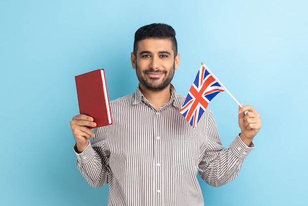 Attractive young adult man holding book and British flag education abroad looking at camera