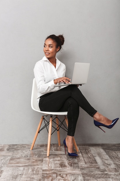 Attraente giovane africano donna d'affari che indossa la camicia seduta su una sedia isolata, lavorando sul computer portatile