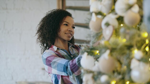 Attractive young African woman decorating Christmas tree at home preparing for Xmas celebration