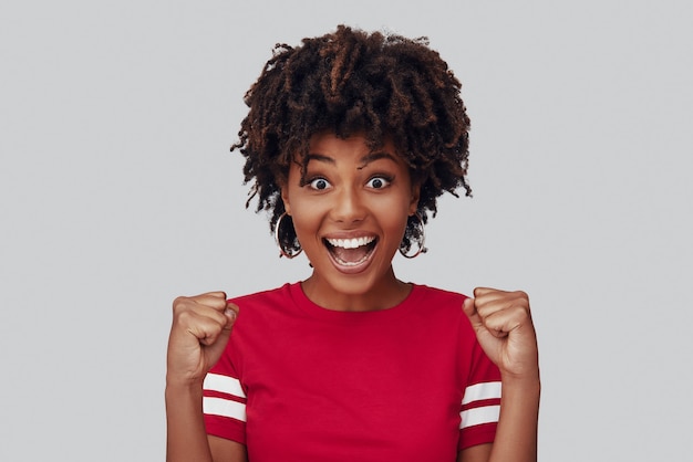 Attractive young African woman looking at camera with smile and gesturing while standing against grey background