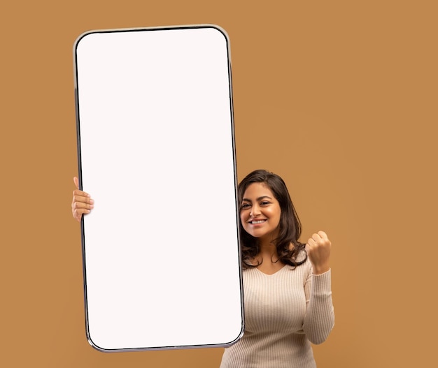 Привлекательная молодая арабская женщина жестикулирует да, держа большой мобильный телефон с пустым экраном над бежевым