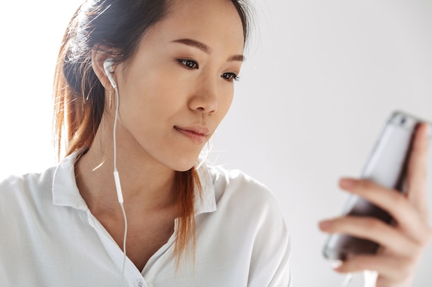 Attractive young asian businesswoman sitting at the office, holding mobile phone, wearing earphones