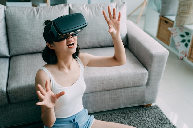 attractive young asian chinese woman in virtual reality headset sitting on floor in house living room. girl in sleepwear wearing vr glasses putting hands in air relax on carpet in apartment indoors