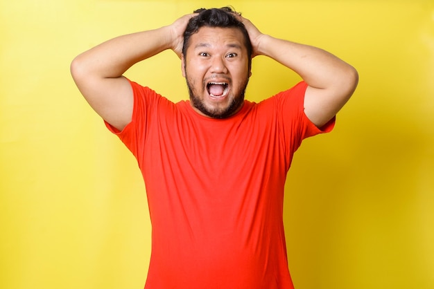 Attractive young Asian fat man in red tshirt with shiny teeth shouting touching head isolated ov