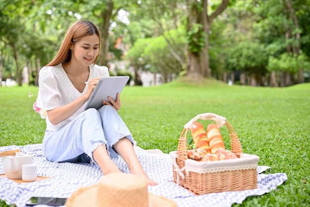 魅力的な若いアジアの女性は、タブレットを使用して、公園でピクニックしながらリラックスします