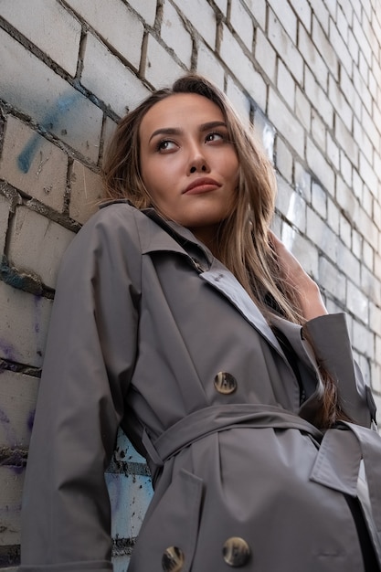 Attractive young asian model in gray trench coat standing near wall outdoors