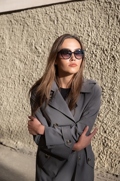 Attractive young asian model in gray trench coat standing near wall outdoors