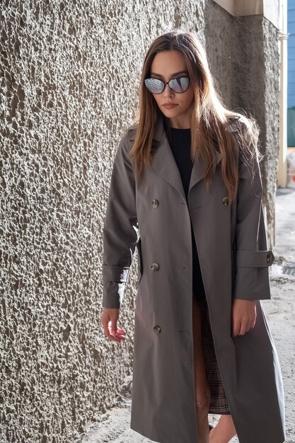 야외 벽 근처에 서 있는 회색 트렌치 코트를 입은 매력적인 젊은 아시아 모델