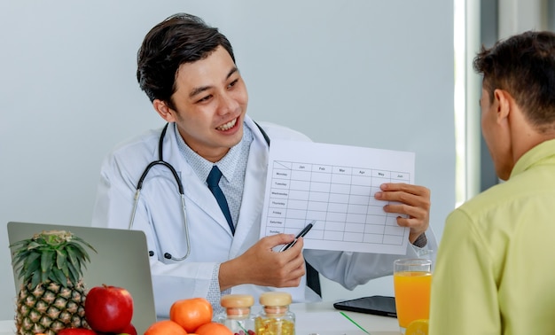 attractive young asian nutritionist doctor wearing stethoscope explaining weekly eating plan with patient. fruits and nutritions and laptop on the table.