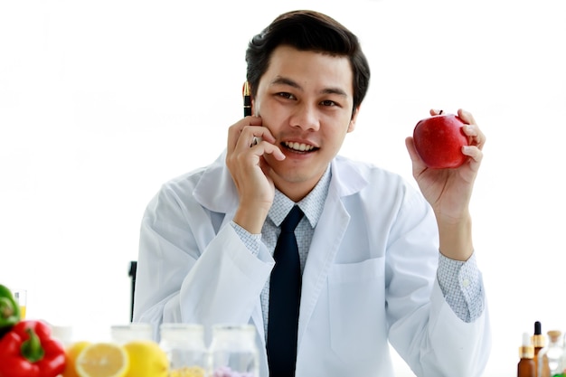 Attractive young Asian nutritionist doctor wearing white lab coat and stethoscope holding red apple up smiling with another hand holding pen. White background, isolated.