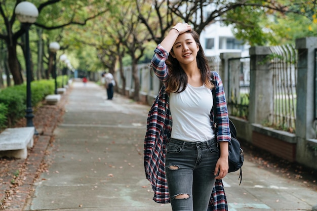 Attraente giovane donna asiatica che gode del tempo fuori nel parco con il sole. studentessa universitaria che va all'università cammina veloce con il vento che soffia e si muove i capelli portando lo zaino all'aperto.