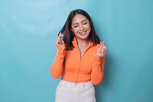 Attractive young Asian woman feels happy and romantic shapes heart gesture expresses tender feelings wears casual orange top against blue background People affection and care concept